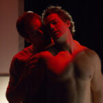 two white men bathed in red light and shadow, one is embracing the other from behind