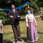 Four people in 19th century clothing stand outdoors, in front of a bright blue and yellow banner.
