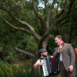 Two men in 19th century clothing stand in front of a green tree. One is playing an accordion.