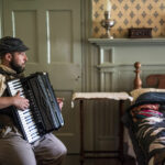 A man dressed in 19th century clothing sits in a bedroom playing an accordion.