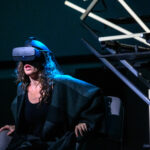 A woman shrouded in black wears a black VR headset. She sits in a chair, but grasps the armrests as if about to stand up.
