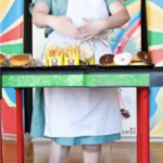 A white woman in a light green cafeteria worker uniform stands behind a counter covered in french fries, hamburgers, and donuts.