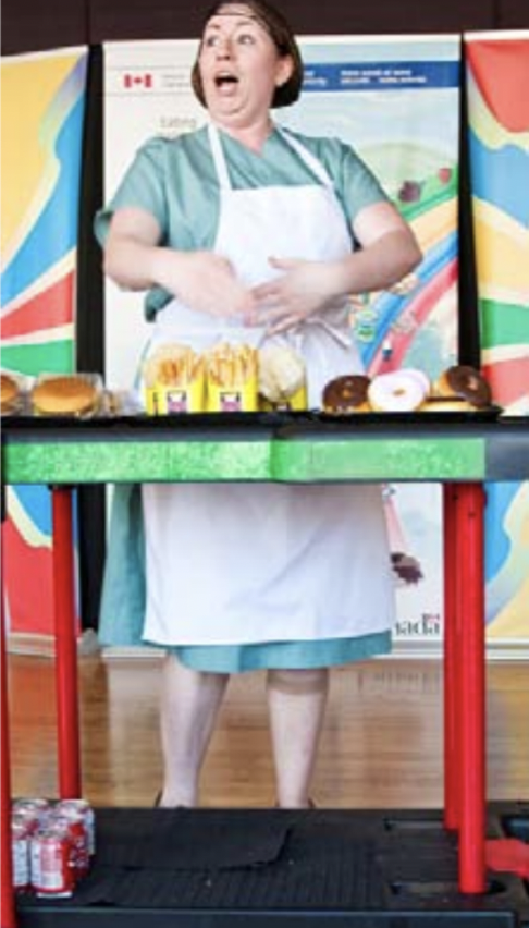A white woman in a light green cafeteria worker uniform stands behind a counter covered in french fries, hamburgers, and donuts.