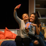 Two young women sit on a bed, taking a selfie posing with a book titled 