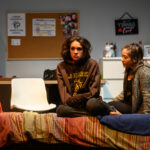 A young woman sits on a bed in a dorm room, dressed in a brown hoodie and with disheveled hair and a dejected expression. Another young woman sits next to her, concerned.