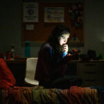 A young woman sits on a bed in a dark dorm room, her face lit by the cell phone she's looking at.