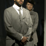 A black man in a grey suit stands in the foreground, while a white woman wearing a grey dress and black hat stands in the background looking at him.