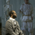A Black man wearing a grey wool suit sits and looks off to the right. In the background, dimly lit, is a skeleton and a man wearing an outfit of crumpled white fabric.