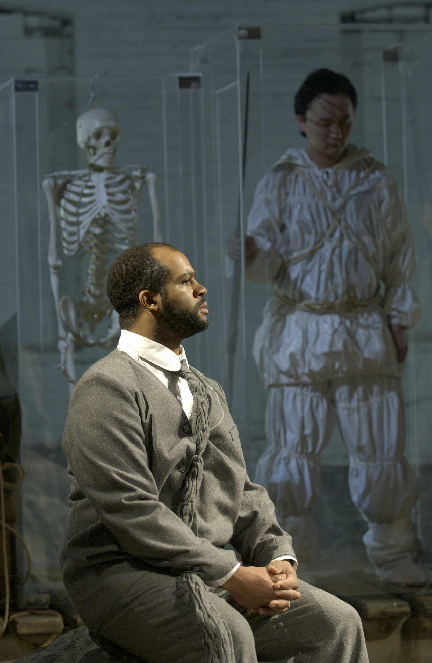 A Black man wearing a grey wool suit sits and looks off to the right. In the background, dimly lit, is a skeleton and a man wearing an outfit of crumpled white fabric.