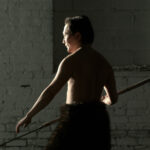 A man stands, lit from behind, mostly in shadow. He is shirtless, wearing bulky black trousers and carrying a spear in one hand.