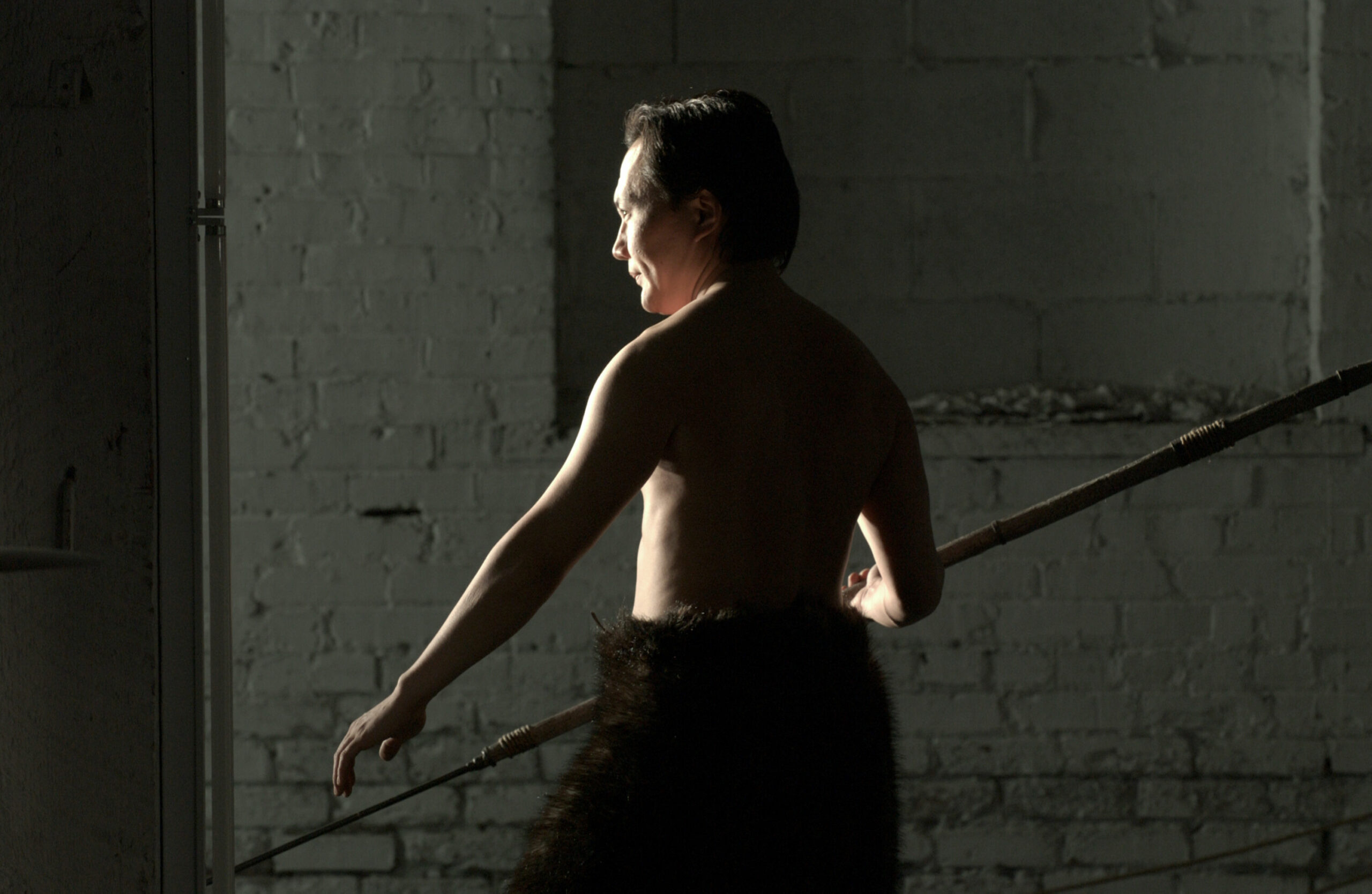 A man stands, lit from behind, mostly in shadow. He is shirtless, wearing bulky black trousers and carrying a spear in one hand.