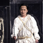 An East Asian man wearing an outfit made of crumpled white fabric and light beige rope stands, holding a spear, inside a rectangular clear glass case. Beside him is a skeleton, in a similar museum-style glass case.