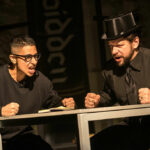 Two people dressed all in black sit at a table, fists clenched on the table. The person on the right wears a black top hat, the person on the left wears round glasses with black frames.