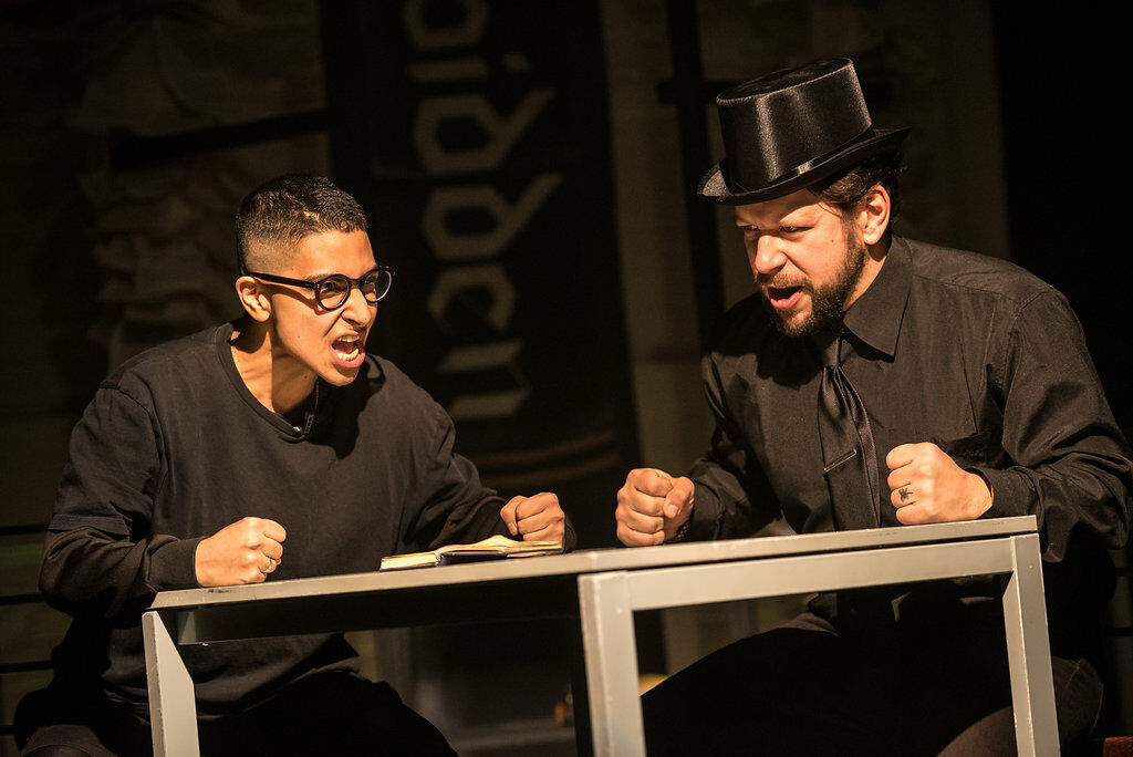 Two people dressed all in black sit at a table, fists clenched on the table. The person on the right wears a black top hat, the person on the left wears round glasses with black frames.