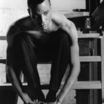 A black and white photo of a Black man, sitting shirtless on a staircase.