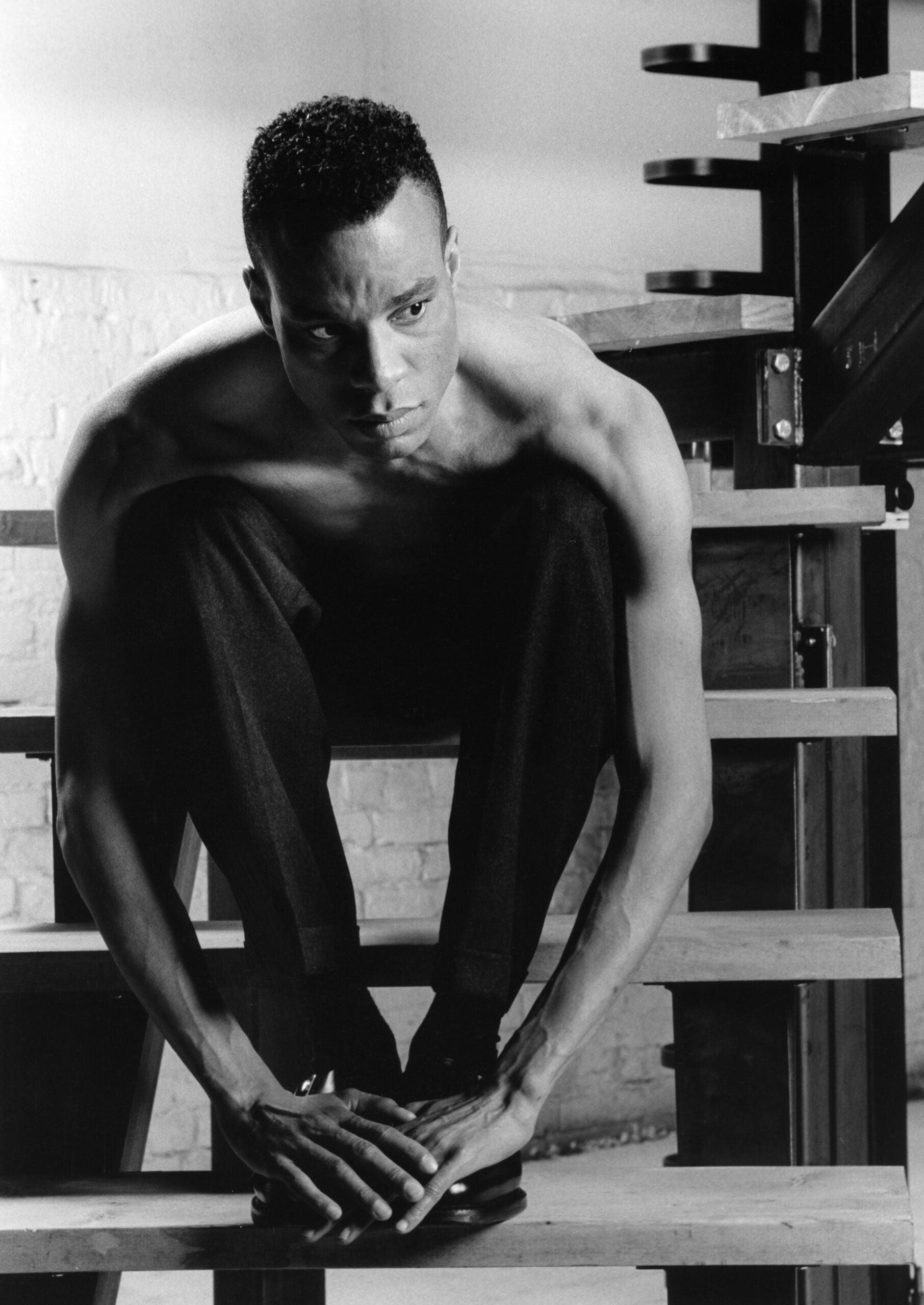 A black and white photo of a Black man, sitting shirtless on a staircase.