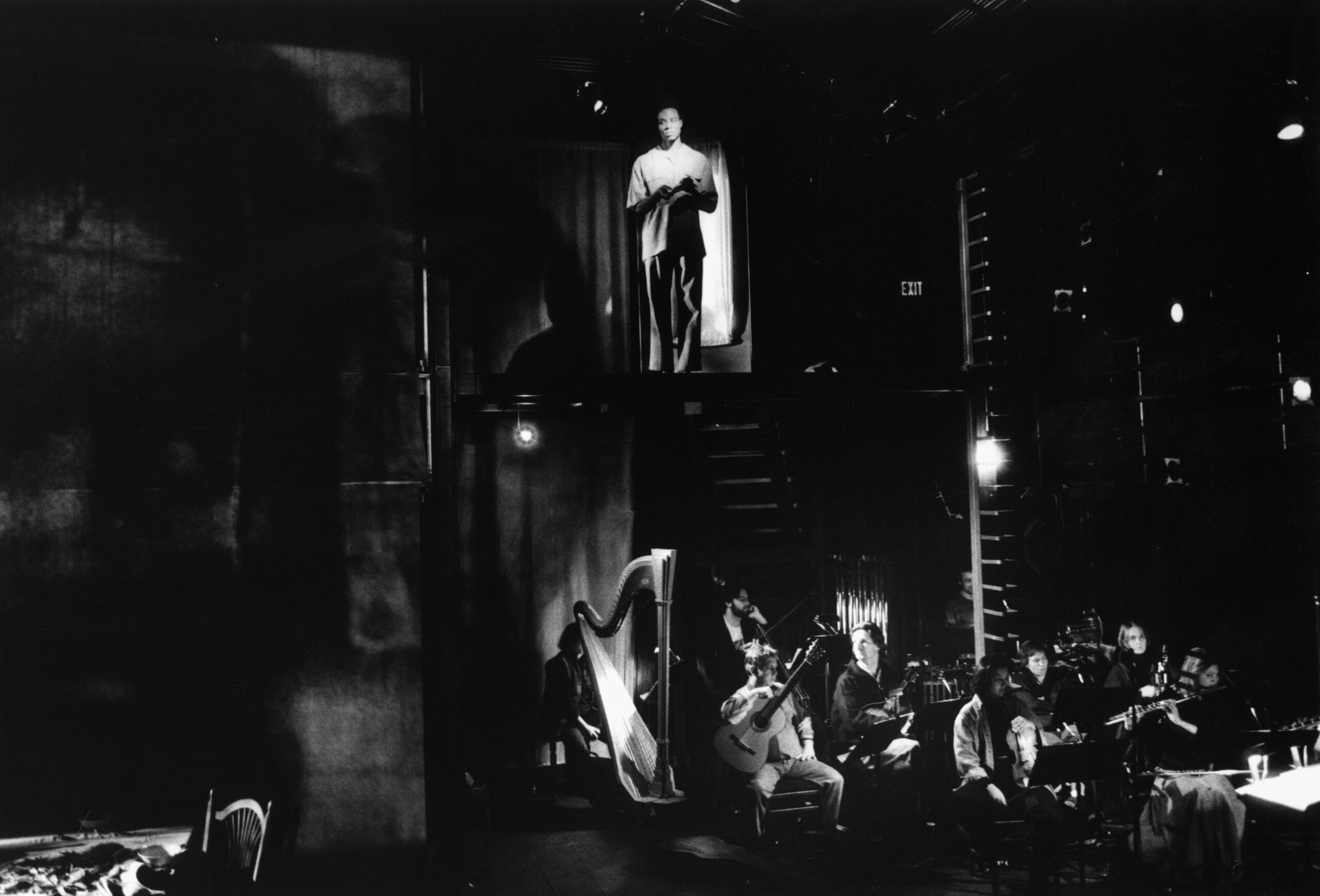 A black and white photo of a Black man standing on the second level of a stage. Underneath on the first level is an orchestra.