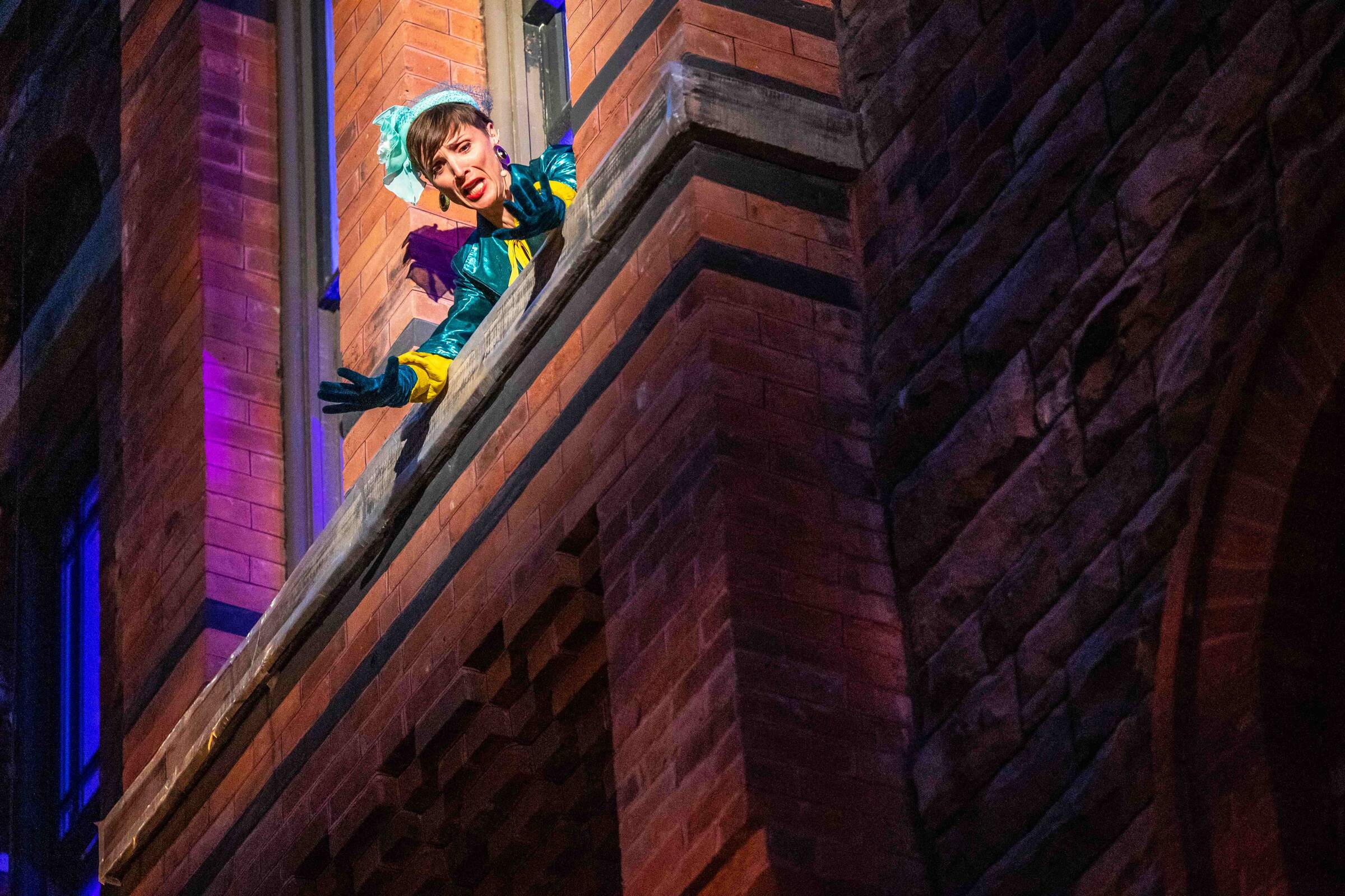A woman wearing a bright teal outfit leans out of a window in a brick wall.