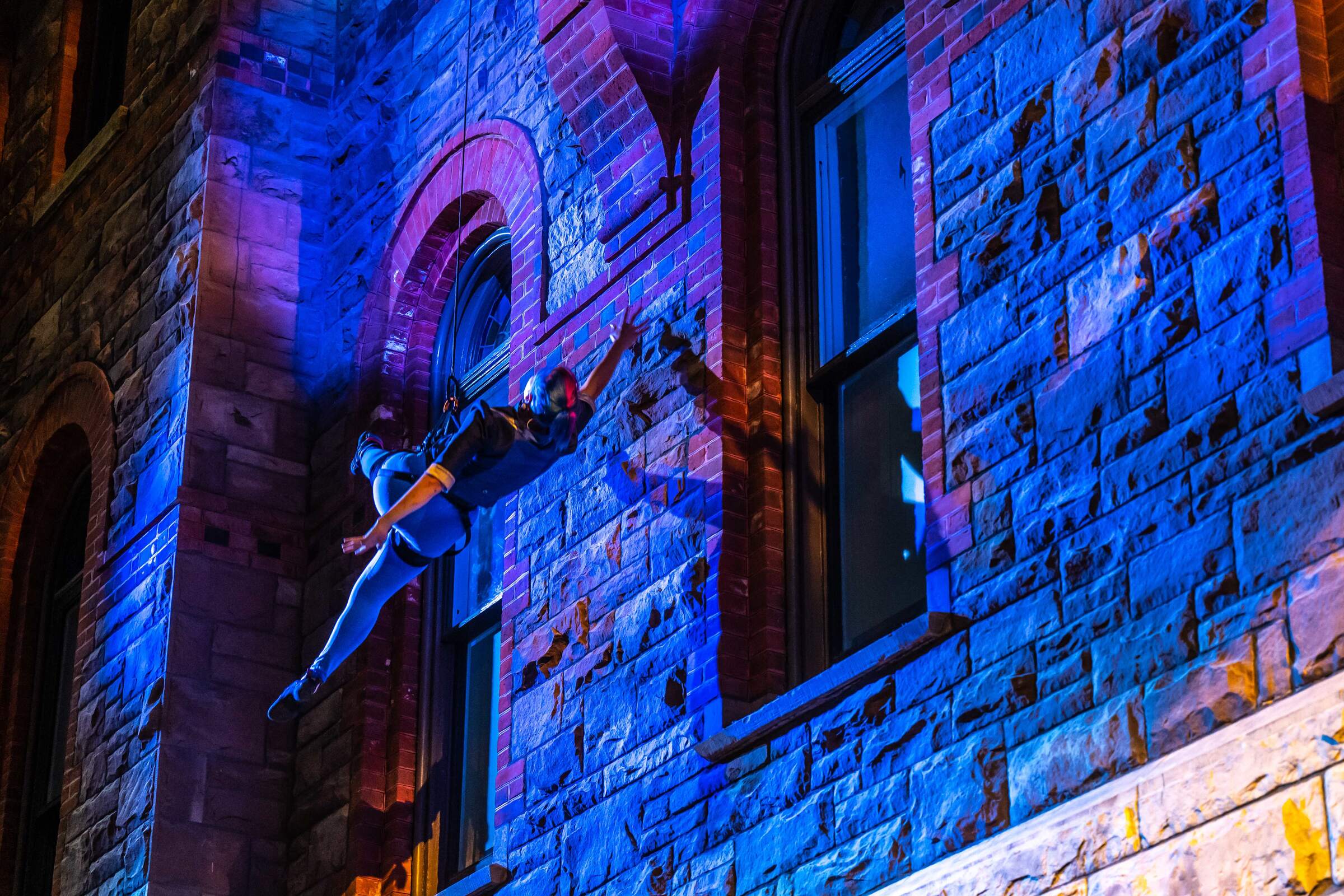 A woman hangs from a climbing harness, suspended next to a brick wall. The entire scene is bathed in dark blue light.