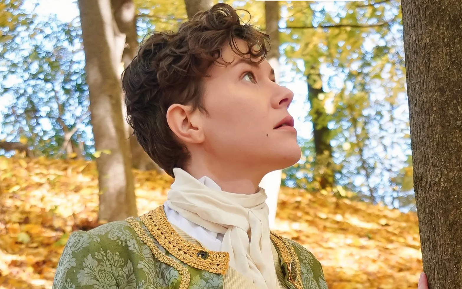 A white genderqueer person with short brown hair looks up to the right. They are wearing fancy 18th century menswear, standing in front of a background of autumn leaves and trees.