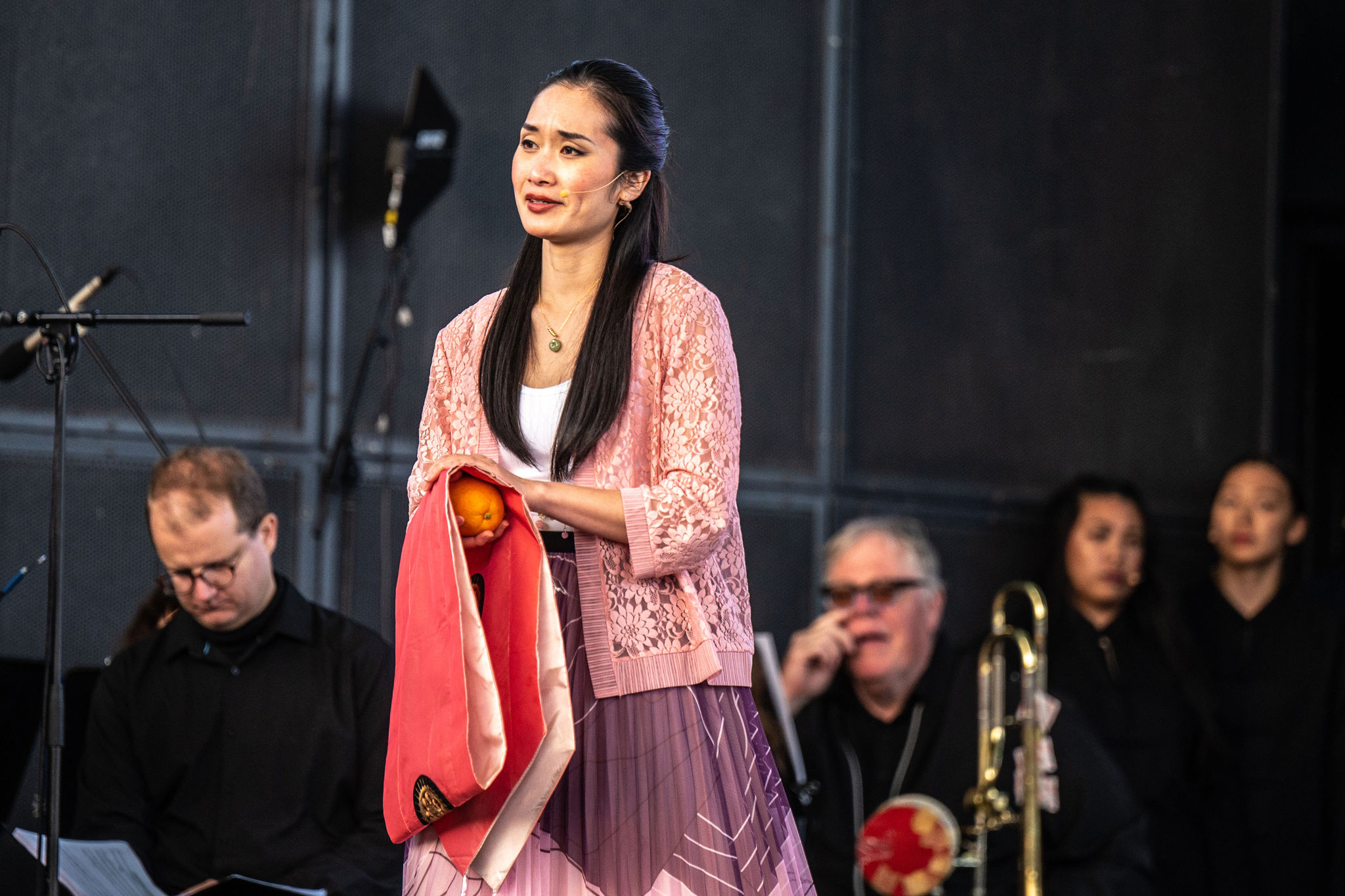 A woman stands in front of an orchestra, wearing a pink lace jacket. She holds a piece of red fabric folded over one arm, and an orange in her hand.