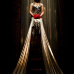 In a dark room, a woman sits on a platform wearing a long silver gown that drapes down below the platform. She holds a bundle of red fabric.