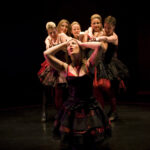 In front of a black backdrop, a woman in a red dress raises her hands to her head in distress. Behind her a group of five women, also dressed in red, reaches towards her.